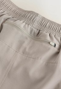 Pantalon beige avec ceinture élastique, poche zippée horizontale et détails de surpiqûres visibles sur un tissu lisse.