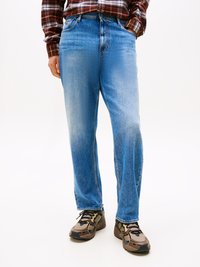 Man wearing blue denim jeans, brown plaid shirt, and brown sneakers standing with one hand in pocket.