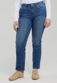 Osoba ubrana w niebieskie jeansy o prostych nogawkach, beżowe trampki oraz biały długi rękaw stojąca na jednolitym tle.