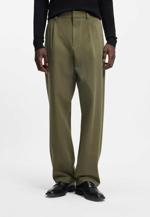 Mężczyzna, model, ubrany w luźne oliwkowozielone spodnie, czarną koszulkę z długim rękawem oraz czarne skórzane buty, stoi na tle jednolitego tła.