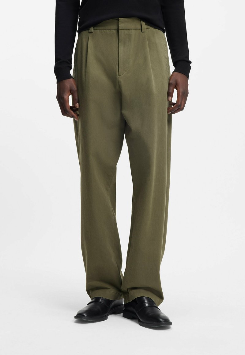 Homme mannequin portant un pantalon ample vert olive, un haut à manches longues noir et des chaussures en cuir noires, debout devant un fond uni.