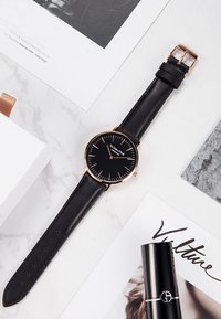 Reloj de cuero negro con una esfera circular negra, acentos en oro rosa y marcadores de horas delgados. Cuenta con una correa de cuero negro con un hebilla de metal.