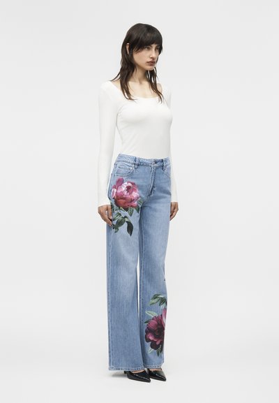 Žena oblečená v bielom top s dlhým rukávom a svetlomodrých širokých džínsach s veľkými ružovými kvetinovými vzormi na ľavom stehne a pravom lýtku, čierne lodičky.