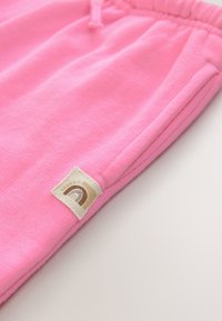 Pantalon de survêtement rose doux avec une taille élastique, une poche latérale et une petite étiquette beige avec un motif arc-en-ciel marron et beige.