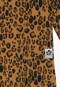 Hladká látka s leopardím vzorem v teplých hnědých a černých odstínech, která má náprsní kapsu a bílou štítkovou nálepku s návrhem pandy a černými akcenty.