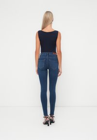 Jeans ajustados en denim azul oscuro con bolsillos traseros y una etiqueta de cuero en la cintura; combinados con una blusa sin mangas negra y tacones de tiras.