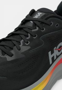 Shoe desportivo preto com parte superior em malha, bico arredondado e acolchoamento espesso. Apresenta detalhes em cinza e laranja, atacadores texturizados e detalhe do logótipo.
