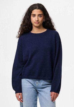 Granatowy sweter z miękkiego materiału, z okrągłym dekoltem i długimi rękawami, zestawiony z jasnoniebieskimi dżinsami. Krótki krój.