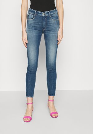 Femme portant un jean skinny bleu taille mi-haute avec un effet délavé et des talons ouverts rose à bride à la cheville, debout sur un fond blanc uni.