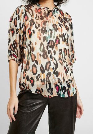 Kvinde iført en knappet transparent bluse med farverigt leopardprint og trekvart puffede ærmer kombineret med sorte bukser.