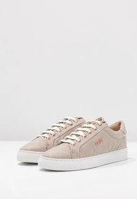 Baskets basses beige avec des lacets blancs, semelles en caoutchouc blanches, motif matelassé et logo métallique "JOOP!" sur le côté.