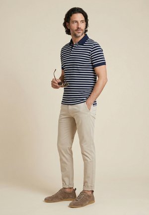 Man draagt een marineblauw-wit gestreept poloshirt, beige broek en bruine sneakers, houdt een zonnebril vast met één hand in zijn zak.