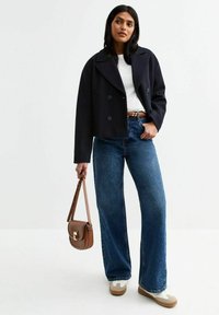 Chaqueta navy corta con gran solapa, botones dobles; camiseta blanca, jeans azules de cintura alta y bolso cruzado marrón con herrajes dorados.