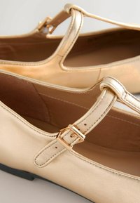Goldene Ballerinas mit T-Riemen-Design, ausgestattet mit einer Metallschnalle und glattem, glänzendem Leder-ähnlichem Material. Flache schwarze Sohle.