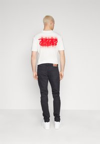 Vit bomullströja med en stor röd grafisk logotyp på baksidan, bärs med svarta jeans och vita sneakers. Korta ärmar, rund hals.