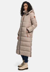 Beige long puffer coat with a hood, featuring quilted sections, front zipper, and orange accents. Worn over blue jeans and cream boots.