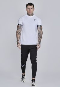 Hvid tætsiddende atletisk skjorte med sorte detaljer på ærmerne, parret med sorte tapered bukser med hvid logo detalje. Hvide sneakers fuldender looket.