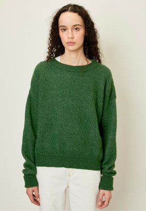 Groene oversized gebreide trui met een geribbelde zoom en mouwen. Zachte textuur, brede halslijn en verlaagde schouders. Gemaakt over een witte broek.