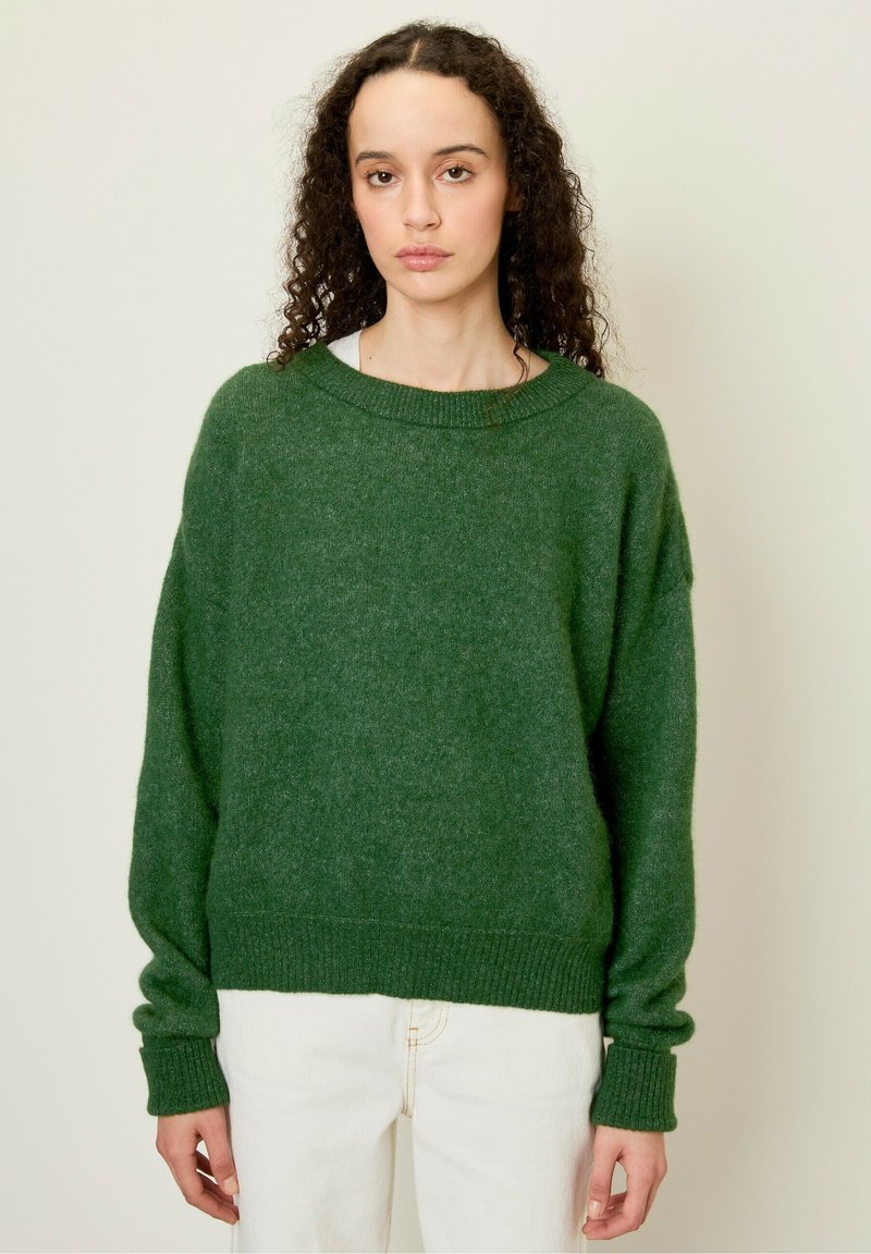 Maglione verde in maglia oversize con orlo e polsini a coste. Tessuto morbido, scollatura ampia e spalle cadenti. Indossato sopra pantaloni bianchi.