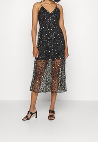 Robe noire à imprimé léopard avec un décolleté en V, des bretelles spaghetti réglables et une superposition transparente jusqu'aux chevilles. Portée avec des sandales à talons noires.