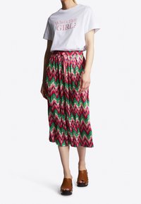 T-shirt blanc avec le texte "Who's that GIRL?", associé à une jupe plissée à motif zigzag multicolore. Des sabots à talons bruns complètent la tenue.