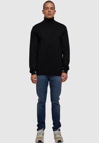 Svart turtleneck-genser laget av myk stoff, parret med blå denimjeans og beige sneakers. Modellen står med hendene langs siden mot en nøytral bakgrunn.