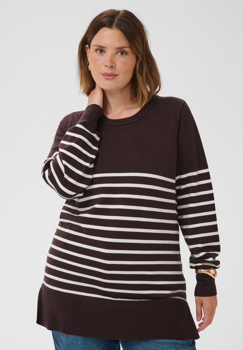 Femme aux cheveux bruns mi-longs portant un pull marron foncé à rayures blanches horizontales et un jean bleu sur un fond uni.