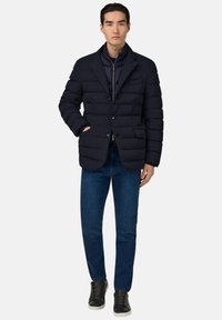 Jūras spēku zila pūkaina jaka ar strukturētu žaketes dizainu, ar pogu aizdari un divām priekšējām kabatām, kombinēta ar tumšiem džinsa biksēm un melniem sporta apaviem.