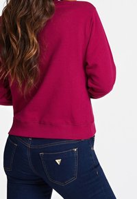 Fuchsia sweatshirt met lange mouwen en een afgeronde zoom, gecombineerd met donkerblauwe jeans met gouden stiksels en een driehoekig logo op de achterzak.