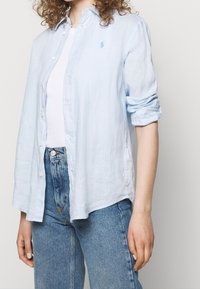 Ljusblå linne-skjorta med knappar fram, långärmad med uppvikta manschetter och en liten broderad logotyp på bröstet. Kombinerad med ljusblå denimjeans.