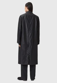 Lange zwarte jas van glanzende, gestructureerde stof met een achterzijde spleet, voorzien van een klassieke kraag en knoopmanchetten, gecombineerd met donkere broek.