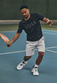 Juodas trumpomis rankovėmis sportinis marškinėliai su "BOSS" logotipu, balti šortai ir juodai balti teniso bateliai. Sportininkas žaidžia tenisą mėlyname korte.