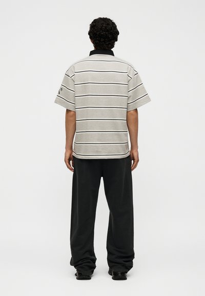 Άνδρας με σγουρά μαλλιά στέκεται με την πλάτη, φορώντας μπεζ πόλο με κοντά μανίκια με μαύρες ρίγες και μαύρο φαρδύ παντελόνι και παπούτσια.