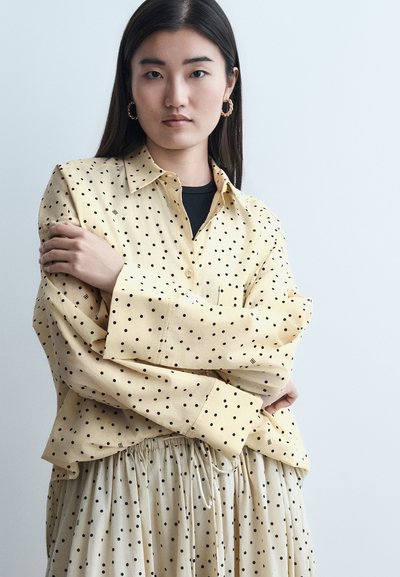 Jeune femme aux longs cheveux foncés portant des boucles d'oreilles créoles dorées, un chemisier jaune à pois et une jupe assortie, debout devant un fond uni.
