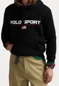 Schwarzer Baumwoll-Hoodie mit "POLO SPORT" in weißen und roten Buchstaben, mit einer kleinen amerikanischen Flagge. Kombiniert mit khakifarbenen Hosen und einem mehrfarbigen karierten Hemd.