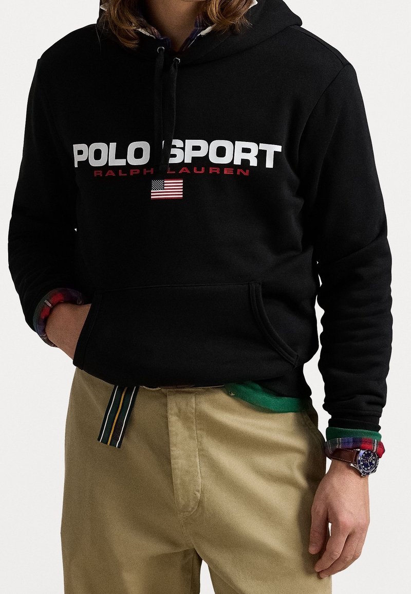 Schwarzer Baumwoll-Hoodie mit "POLO SPORT" in weißen und roten Buchstaben, mit einer kleinen amerikanischen Flagge. Kombiniert mit khakifarbenen Hosen und einem mehrfarbigen karierten Hemd.