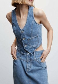 Denim vest in light blue, featuring a deep v-neck, five buttons, and two front pockets with a fitted design. Worn with matching denim skirt.