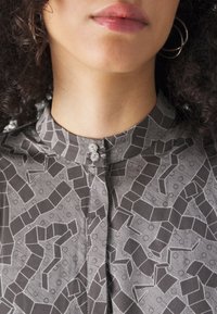 Hallitatud hall blouse geomeetriliste mustritega, krae nuppe detailiga ja siidise tekstuuriga. Aktsentideks on kaks dekoratiivset nuppu kaeluses.
