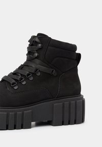 Botte noire montante avec semelle plateforme épaisse rainurée et larges lacets noirs passant dans des œillets métalliques sur fond uni.
