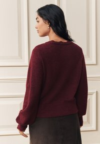 Bordeauxroter Strickpullover mit strukturierter Oberfläche, mit einem gewellten Ausschnitt und langen Ärmeln, kombiniert mit einem dunklen Rock.