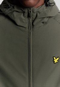 Giacca verde con zip e cappuccio, caratterizzata da un logo di aquila gialla sul petto e da lacci elastici sul cappuccio per la regolazione.