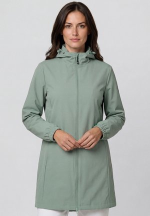 Frau trägt eine hellgrüne Kapuzenjacke mit Frontreißverschluss und elastischen Bündchen, steht vor einem einfarbigen grauen Hintergrund.