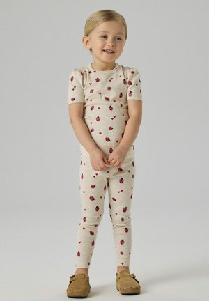 Lachend jong kind staand, gekleed in crèmekleurige outfit met lieveheersbeestje- en hartafdrukken, en lichtbruine instapschoenen tegen een effen achtergrond.