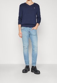 Suéter de cuello en V navy, jeans ajustados de color azul claro y botas gruesas negras. El suéter cuenta con puños acanalados y un pequeño logo en el pecho.
