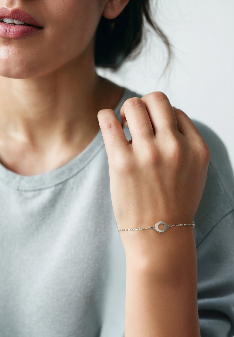 Bracciale d'argento con un ciondolo esagonale, catena fine, indossato su un polso, abbinato a una maglietta grigia chiara a collo rotondo con texture.