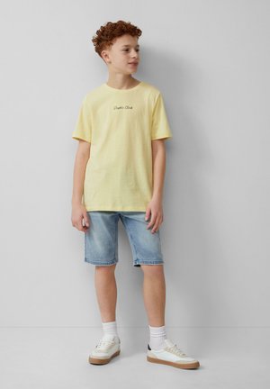 Geel T-shirt met korte mouwen en zwarte tekst, gecombineerd met lichtblauwe denimshorts en witte sneakers met beige zolen en marineblauwe accenten.