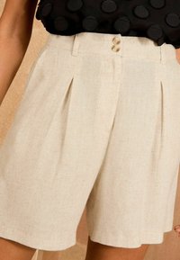 Beige vászon rövidnadrág pliszékkel, két elülső gombbal. Az anyag finoman textúrázott, egyenes vonalú és szegéllyel ellátott.