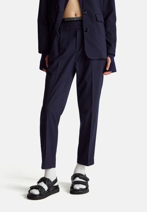 Marineblauwe streeppantalons met een getailleerde pasvorm, voorzien van een rekbare tailleband en opgerolde boorden. In combinatie met zwarte sandalen en witte sokken.