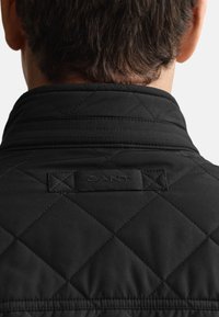 Czarna pikowana kurtka z wysokim kołnierzem i metką marki GANT z tyłu. Materiały o fakturze z przeszywanym wzorem w kształcie diamentu na całej powierzchni.