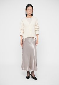 Cream cable-knit cardigan with a V-neck, paired with a silver satin midi skirt and black pointed-toe heels, standing against a plain background.